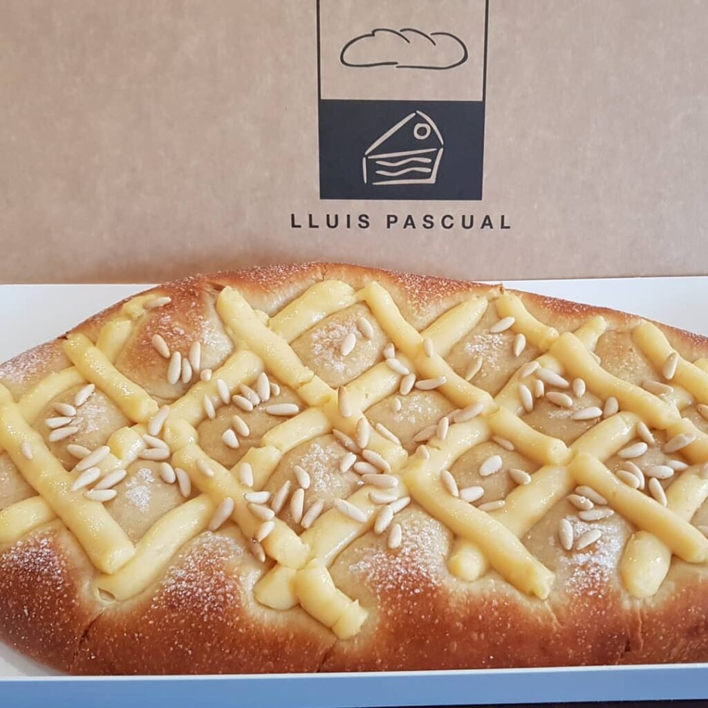 Coca de brioche esponjosa, decorada con crema pastelera y piñones, elaborada artesanalmente en la pastelería Lluis Pascual. Ideal para celebraciones.