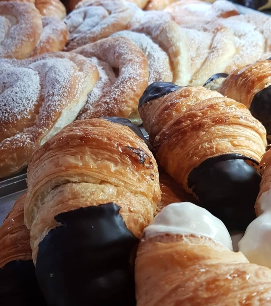 Primer plano de croissants rellenos de chocolate blanco y negro, con cobertura de chocolate, acompañados de ensaimadas espolvoreadas con azúcar glas. Bollería artesanal ideal para desayunos.