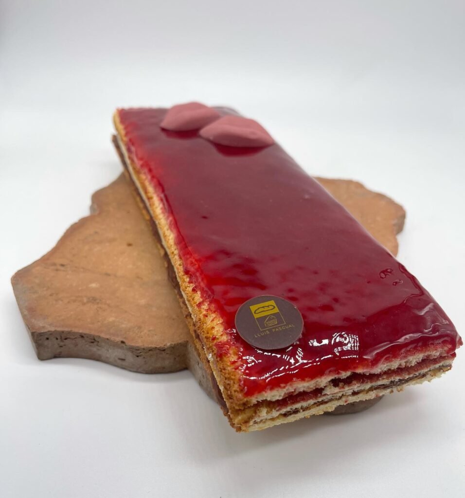 Pastel rectangular de frambuesa y chocolate, con una capa brillante de frambuesa, decorado con detalles de chocolate y sello de Lluis Pascual. Elegante y delicioso.
