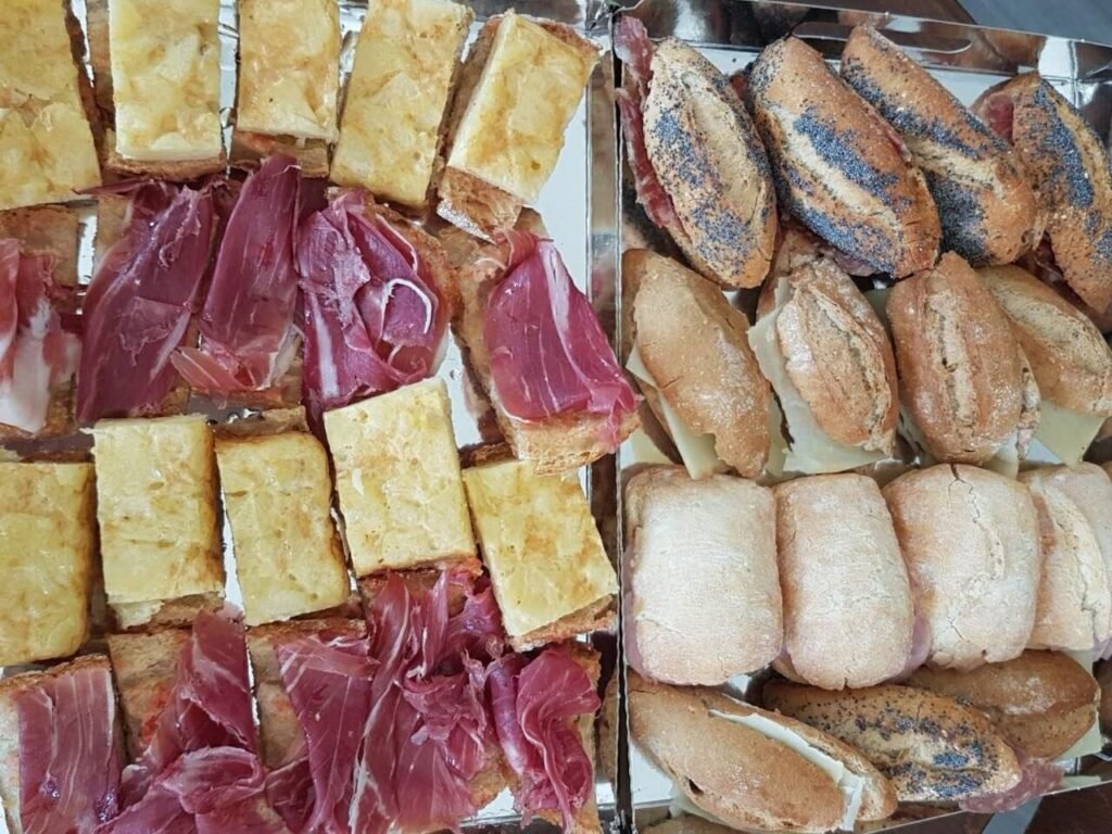 Bandeja con una selección de bocadillos: mini bocadillos de pan rústico con jamón serrano y queso, y tostadas con tortilla de patata y jamón ibérico. Perfecto para catering o eventos.